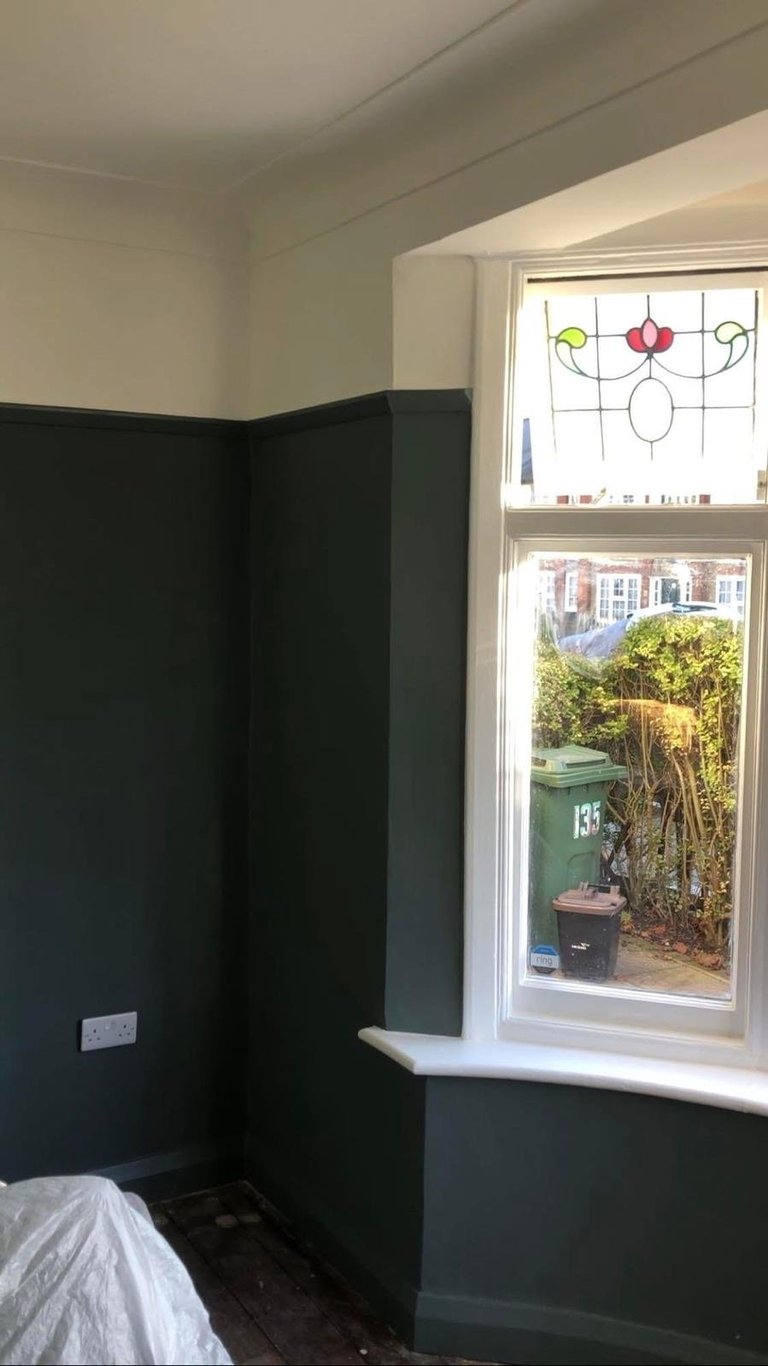 Room corner with dark lower half walls, cream upper walls, and window with stained glass decoration overlooking a garden