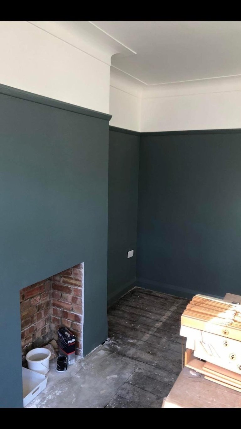 Interior room under renovation with dark teal walls, exposed brick fireplace opening, concrete floor, and wooden furniture stacked on the right