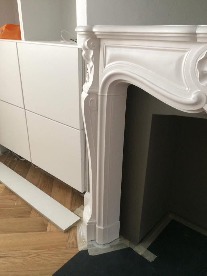 White fireplace mantel with decorative trim next to a white cabinet on wooden floor