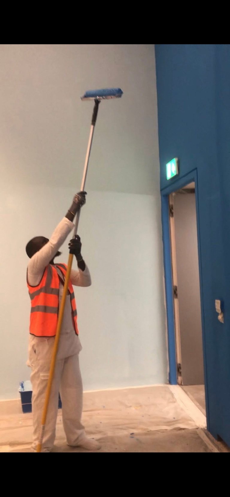 Worker in orange safety vest painting a white wall with roller on an extended pole indoors