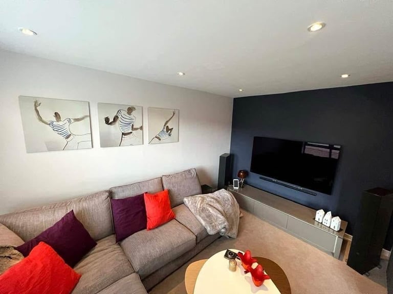 Modern living room with beige L-shaped sofa, colorful cushions, wall art, flat-screen TV on dark accent wall, and round coffee table