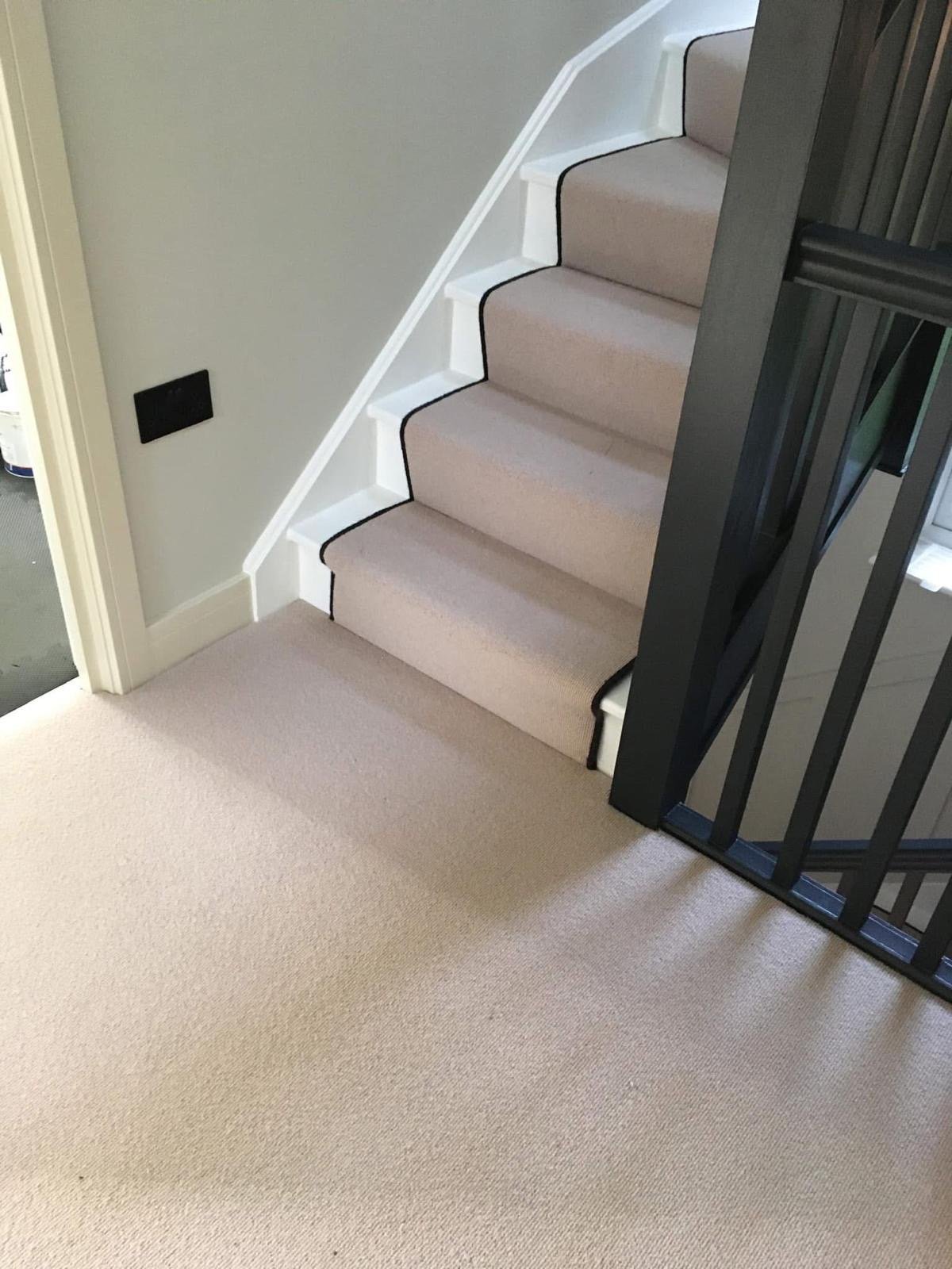 Staircase with beige carpet and black binding, white handrail, viewed from landing with dark gate visible